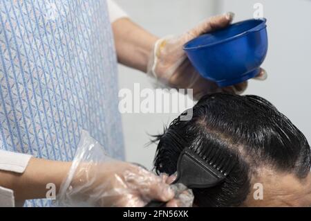 Friseur mit Einweg-Plastikhandschuhen, die die Haarwurzeln einer Frau färben Mit einer Haarbürste Stockfoto