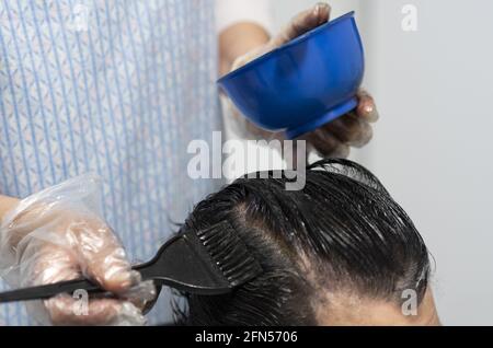 Friseur mit Einweg-Plastikhandschuhen, die die Haarwurzeln einer Frau färben Mit einer Haarbürste Stockfoto