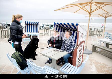 Norderney, Deutschland. Mai 2021. Die Kellnerin Nina (l.) bedient Anet und Christoph aus der Nähe von Oldenburg, die mit ihrem Hund Murphy in einer Strandliege vor dem Surfcafe an der Strandpromenade sitzen. In Niedersachsen dürfen Hotels, Ferienwohnungen und andere Unterkünfte in Regionen mit einer siebentägigen Inzidenz von weniger als 100 Tagen wieder Gäste empfangen. Quelle: Hauke-Christian Dittrich/dpa/Alamy Live News Stockfoto