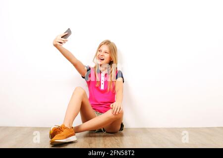 Schönes Teenager-Mädchen, das auf Holzboden sitzt und Selfie-Aufnahmen auf ihrem Handy gemacht hat. Lässige junge Frau in gelben Sneakers fotografiert sich selbst auf SM Stockfoto