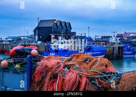 Whitstable Harbour, Kent, England, Großbritannien - farbenfrohe Fischernetze und Bojen am Kai mit Fischerboot und Lagerhäusern im Hintergrund Stockfoto