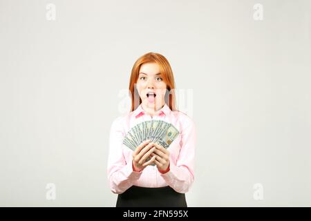 Junge, schöne Rothaarige Frau, die ihr Gesicht mit einer Handvoll Geld bedeckt, das wie ein Fan mehrere hundert-Dollar-Scheine hält. Aufgeregt attraktive weibliche wi Stockfoto