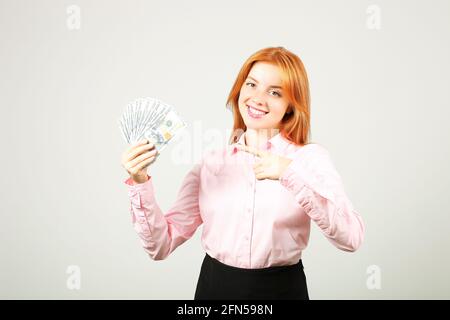 Junge, schöne Rothaarige Frau, die mit dem Zeigefinger auf eine Handvoll Geld zeigt, das wie ein Fan einen Hundert-Dollar-Scheine hält. Aufgeregte attraktive Hündin Stockfoto