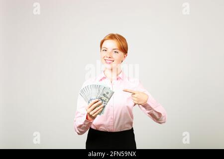 Junge, schöne Rothaarige Frau, die mit dem Zeigefinger auf eine Handvoll Geld zeigt, das wie ein Fan einen Hundert-Dollar-Scheine hält. Aufgeregte attraktive Hündin Stockfoto