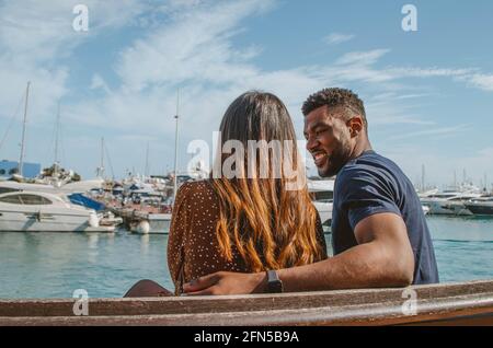 Multiethnisches Paar, das auf einer Bank vor dem sitzt Hafen mit auf dem Wasser schwimmenden Booten Stockfoto
