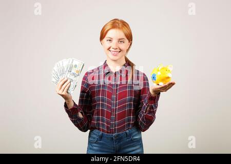 Porträt einer jungen, schönen Rotschopf-Frau im Flanellhemd mit einem Stapel von hundert Dollar Scheinen in einer Hand und einem gelben Sparschwein in einer anderen. Geben Sie oder aus Stockfoto