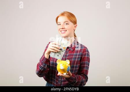 Porträt einer jungen, schönen Rotschopf-Frau im Flanellhemd mit einem Stapel von hundert Dollar Scheinen in einer Hand und einem gelben Sparschwein in einer anderen. Geben Sie oder aus Stockfoto