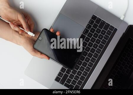 Nahaufnahme der Hände, die ein schwarzes Smartphone halten und ein Kabel über einem Laptop anschließen. Stockfoto