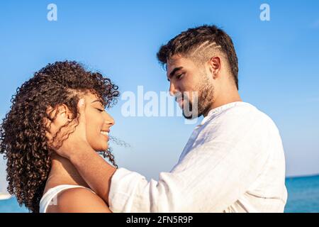 Moment der Zärtlichkeit zwischen jungen, multirassischen Paaren glücklicher Liebhaber, die einander in die Augen schauen, während er lächelnd ihren Kopf in den Händen hält. Bestanden Stockfoto