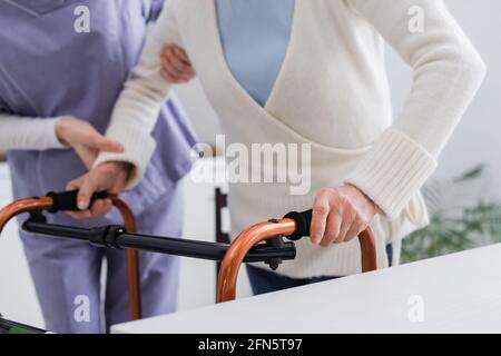 Teilansicht der Krankenschwester, die eine ältere Frau unterstützt, die mit medizinischen Gehern geht, verschwommener Hintergrund Stockfoto