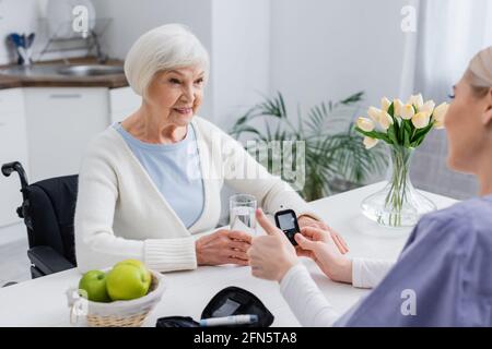 Krankenschwester zeigt Daumen in der Nähe von Glucometer und lächelnde diabetische Frau, verschwommener Vordergrund Stockfoto