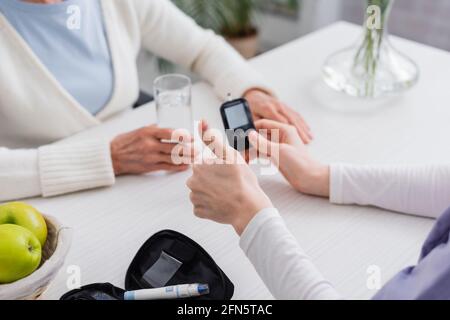 Beschnittene Ansicht eines Sozialarbeiters, der den Daumen in der Nähe des Glukosimeters zeigt Und ältere Diabetikerin Stockfoto