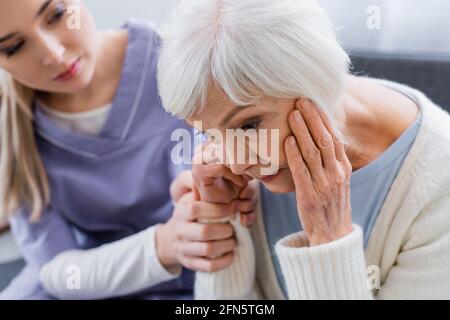 Junge Krankenschwester berührt die Hand einer älteren Frau, leidet an Demenz und sitzt mit gesenktem Kopf, verschwommener Hintergrund Stockfoto