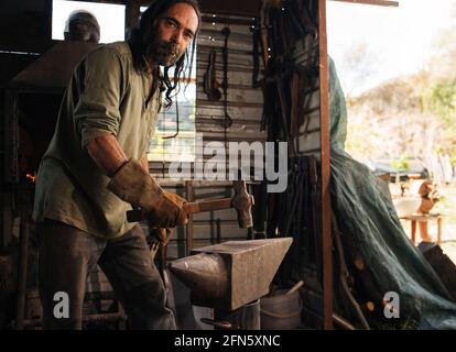 Langhaariger Schmied, der in seiner Werkstatt auf einem Amboss eine Keule zeigt. Stockfoto
