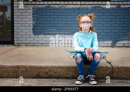 Rotschopf kleines Mädchen sitzt auf einem Bordstein mit Skateboard Blasen Blase. Stockfoto