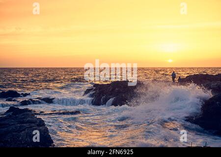 Spritzer von den Wellen, die beim Sonnenuntergang gegen das felsige Ufer stoßen. Stockfoto