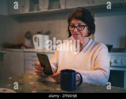 Lächelnde ältere Frau, die in der Küche sitzt und im Internet surft Auf dem Mobiltelefon Stockfoto