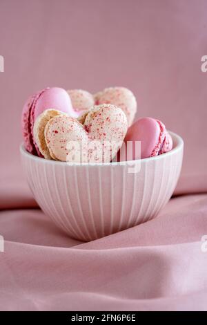 Eine Gruppe herzförmiger französischer Macaronkekse in einer rosa Schale auf einem rosa Stoffhintergrund, Vanille- und Erdbeergeschmack, vertikal, Kopierfläche Stockfoto
