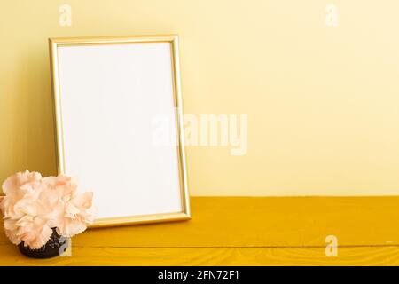 Leerer Bilderrahmen mit Nelkenblumen auf Holztisch. Gelber Hintergrund Stockfoto