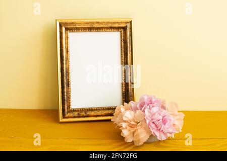 Leerer Bilderrahmen mit Nelkenblumen auf Holztisch. Gelber Hintergrund Stockfoto
