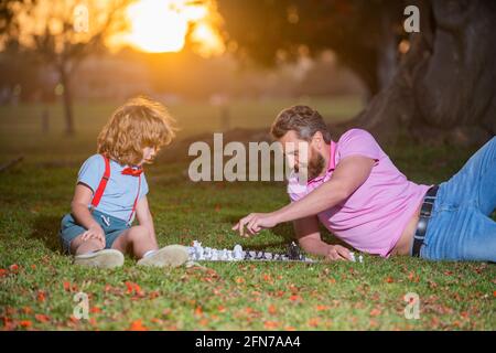 Vater und Sohn spielen Schach und liegen auf dem Rasen im Rasenpark. Vatertag, Liebesfamilie, Elternschaft, Kindheitskonzept. Aktive Menschen, die Spaß haben und Stockfoto