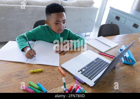 afroamerikanischer Junge im Online-Schulunterricht, mit Laptop und Schreiben in seinem Notebook Stockfoto