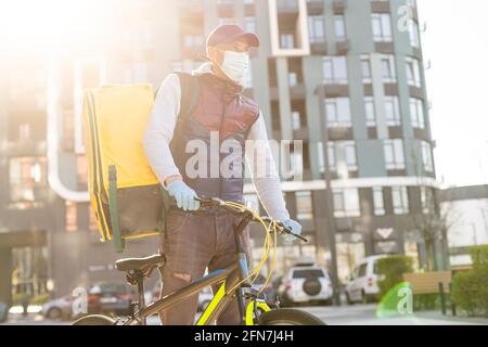 Junger Kurier in medizinischer Maske liefert Lebensmittel mit gelbem Thermalrucksack, fährt mit dem Fahrrad in der Stadt. Konzept für den Lieferservice von Lebensmitteln Stockfoto