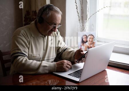 Lächelnder älterer Mann trägt Kopfhörer winken zur Kamera und hat Videoanruf auf dem Laptop, glücklicher älterer Mann mit Kopfhörern sitzt zu Hause auf der Couch und redet mit Modern Stockfoto