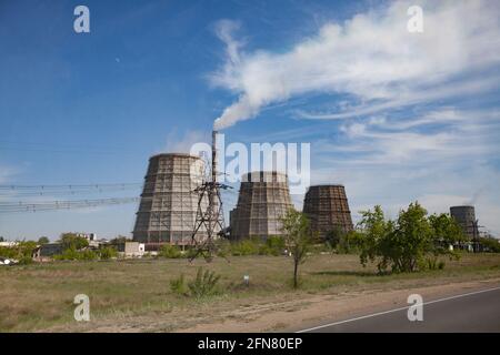 Pavlodar-Wärmekraftwerk. Kühltürme und Rauchschwaden mit weißem Rauch. Blauer Himmel. Grünes Gras, Bäume. Asphaltstraße rechts. Stockfoto