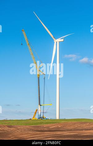 Windturbine im Bau in der Nähe eines Dorfes auf einem landwirtschaftlichen Feld mit klarem blauen Himmel auf dem Hintergrund. Konzept für erneuerbare Energien, grün ökologisch Stockfoto