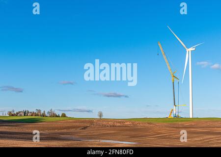 Windturbine im Bau in der Nähe eines Dorfes auf einem landwirtschaftlichen Feld mit klarem blauen Himmel auf dem Hintergrund. Konzept für erneuerbare Energien, grün ökologisch Stockfoto