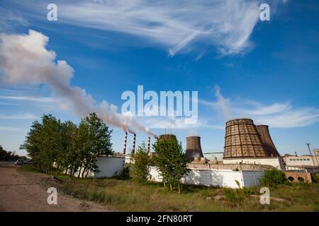 Pavlodar-Wärmekraftwerk. Kühltürme und Rauchschwaden mit weißem Rauch. Grünes Gras, Bäume, blauer Himmel mit Wolken. Stockfoto