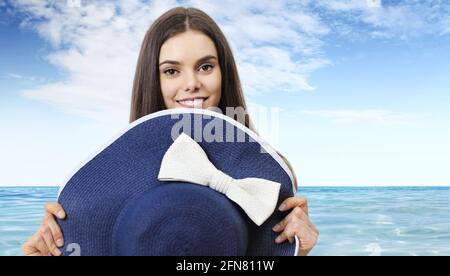 Porträt der lächelnden schönen Brünette junge Frau versteckt sich hinter blau Strohhut isoliert auf Meer und Himmel Hintergrund Stockfoto