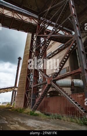 Temirtau, Kasachstan: Veraltete sowjetische Zementfabrik. Durchbrochene Metallstütze des Förderers. Alte Asphaltstraße. Rauchschwaden und grauer wolkig. Stockfoto