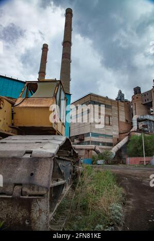 Temirtau, Kasachstan: Veraltete sowjetische Zementfabrik. Alte Bulldozer links auf dem Gras. Rauchschächte, Industriegebäude auf grau bewölktem Himmel. Stockfoto