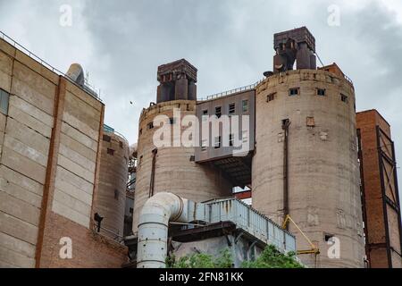 Alte veraltete sowjetische Zementanlage veraltete Zementsilos (Türme). Metallgraues Rohr vor dem Gebäude, Industriegebäude links. Temirtau, Kasachstan. Stockfoto