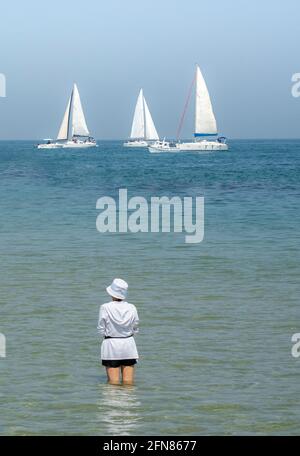 Eine junge Frau in einem weißen Top und Hut, die an einem sonnigen Tag im flachen Meerwasser steht und Segelboote vorbeifahren sieht. Stockfoto