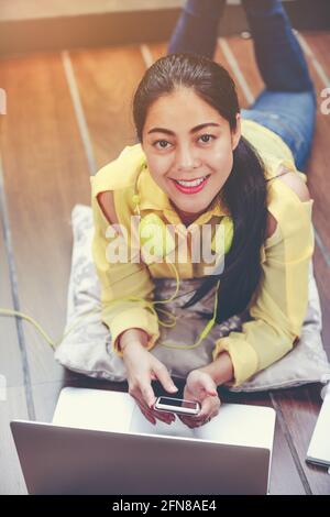 Glückliche asiatische Frau, die lächelt und einen Laptop benutzt, während sie auf dem Boden mit Kopfhörern zur Unterhaltung liegt. Frau, die am Laptop arbeitet und die Kamera anschaut Stockfoto