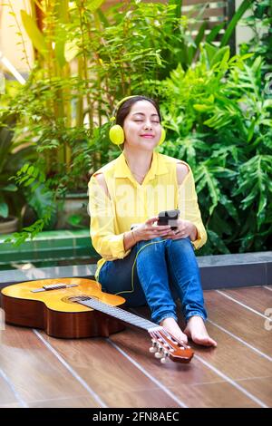 Asiatische Musikerin mit Mobiltelefon mit Kopfhörern für Unterhaltung und akustische Gitarre in der Nähe. Frau genießt das Surfen im Internet auf Smart phon Stockfoto