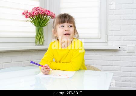 Das kleine Mädchen sitzt an einem Tisch in einem weißen Raum und zeichnet. Stockfoto