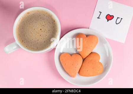 Hausgemachte herzförmige Kekse, eine Tasse Kaffee und eine Notiz mit den Worten, die ich dich liebe, in pastellrosa Farbe. Frühstück am Valentinstag. Valentinstag Stockfoto