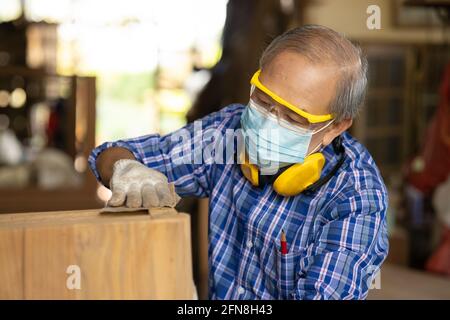 Ältere Arbeiter Holzholzarbeiten Ruhestand Hobby für einen guten Ruhestand, asiatische männliche reifen professionellen Meister der Herstellung von Holzmöbeln mit Gesichtsmaske Protecti Stockfoto