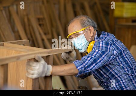 Ältere Arbeiter Holzholzarbeiten Ruhestand Hobby für einen guten Ruhestand, asiatische männliche reifen professionellen Meister der Herstellung von Holzmöbeln mit Gesichtsmaske Protecti Stockfoto