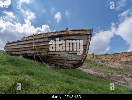 Ein verstörtes hölzernes Fischerboot, das an sonnigen Tagen auf Grasflächen mit blauem Himmel im Hintergrund abgesetzt wurde. Stockfoto