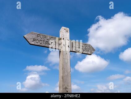 Ein doppeltes öffentliches Wanderwegschild, das in entgegengesetzte Richtungen auf einen Holzpfosten mit blauem Himmel und Wolken im Hintergrund zeigt. Stockfoto