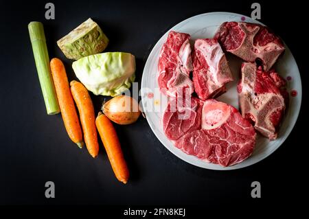Ungekochte Speisen. Rohe Rinderknochen und Gemüse auf schwarzem Hintergrund. Zutaten für die Herstellung von Rinderbrühe. Gesunde Ernährung. Stockfoto
