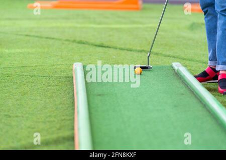 Person spielt ein Spiel von Minigolf auf Kunstrasen in einem niedrigen Winkel abgeschnitten Ansicht ihrer Beine, der Ball und Golfschläger mit Copyspace Stockfoto