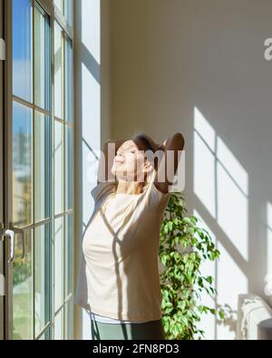 Genießen Sie den neuen Tag. Schöne glückliche reife Frau, die sich am sonnigen Morgen zu Hause mit geschlossenen Augen und Sonnenstrahlen durch das Fenster streckt und lächelt. Optimismus und Glück Konzept Stockfoto