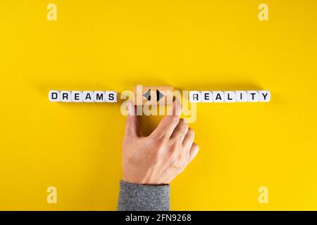 Die Hand hält einen Holzwürfel mit Pfeilsymbol zwischen den Optionen von Träumen oder Realität. Den Träumen zu jagen oder dem Dilemma der Realität zu begegnen. Stockfoto