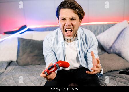 Foto eines gutaussehenden aufgeregt Kerl sitzt auf dem bequemen Sofa, Quarantäne bleiben zu Hause Gamer hält Spielstation Joystick, gute Laune fifa Meisterschaft, unglücklich Stockfoto